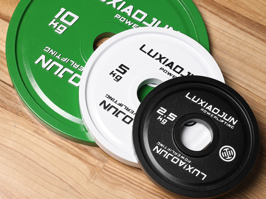 Green 10 kg, white 5 kg and black 2.5 kg LUXIAOJUN powerlifting plates overlapping on a wood floor, showing the painted finish and weight markings